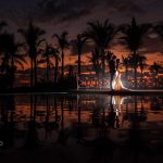 The groom and the bride in formal dress are posing for a sunset shot on the border of a pool where are reflected the palms.