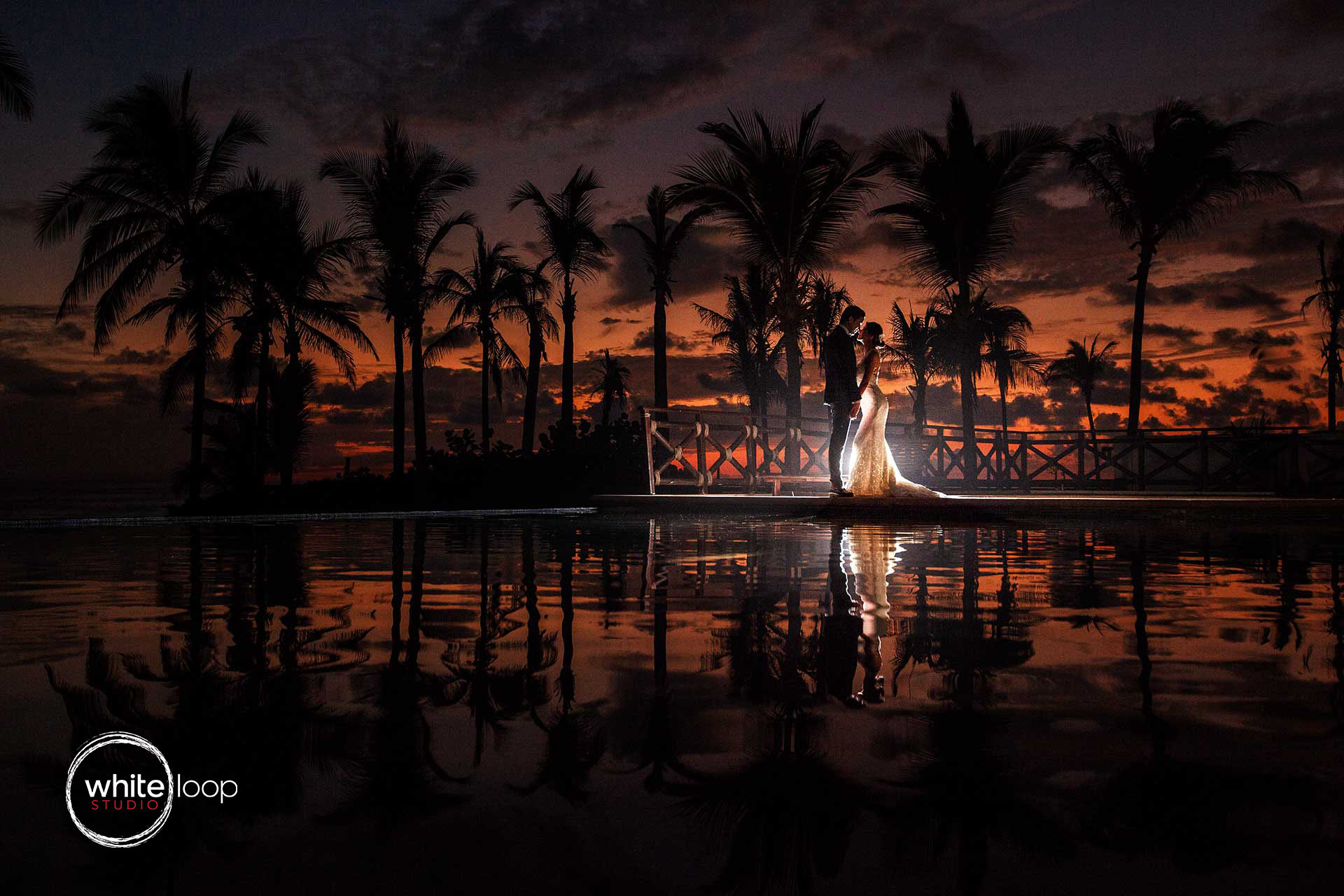 The groom and the bride in formal dress are posing for a sunset shot on the border of a pool where are reflected the palms.