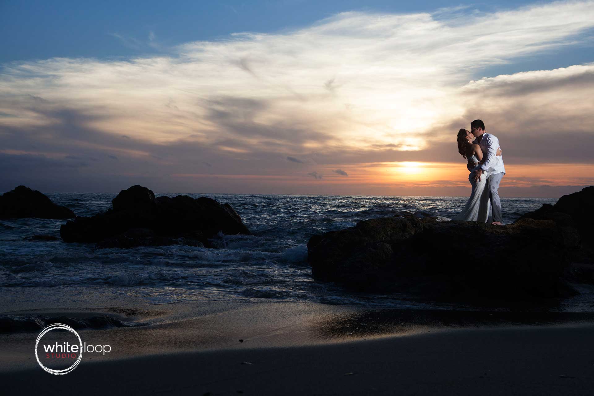 The groom and the bride embrace on top of a rock on the seashore.